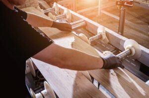 Worker handling wooden planks on a production line, showcasing the manufacturing process of flooring materials with sunlight streaming in.