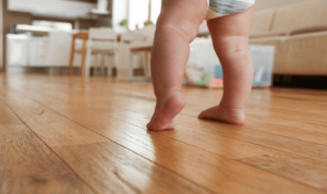 Baby taking first steps on smooth wooden flooring in a cozy living room with blurred furniture in the background.