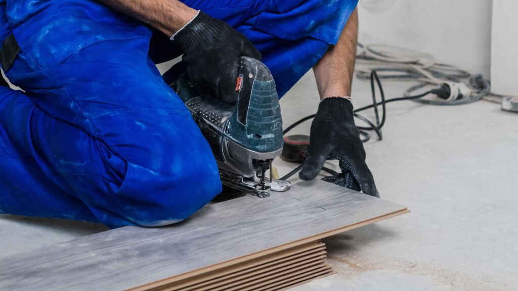 Worker cutting vinyl plank flooring with electric jigsaw tool.