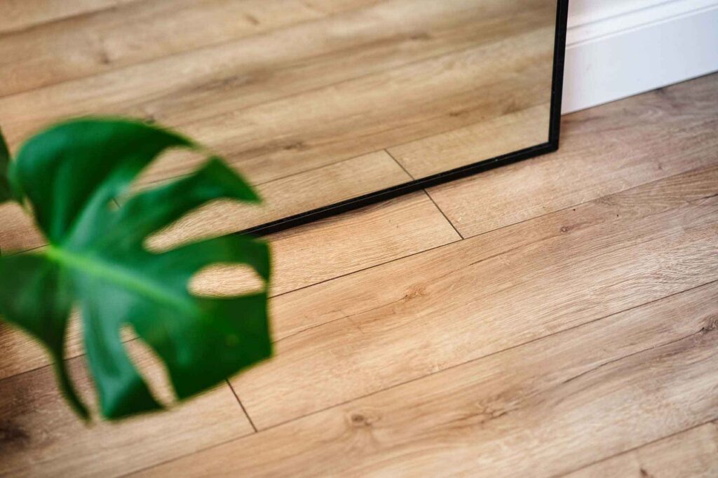 Close-up of natural oak laminate flooring with a large mirror and green plant leaf in the corner.