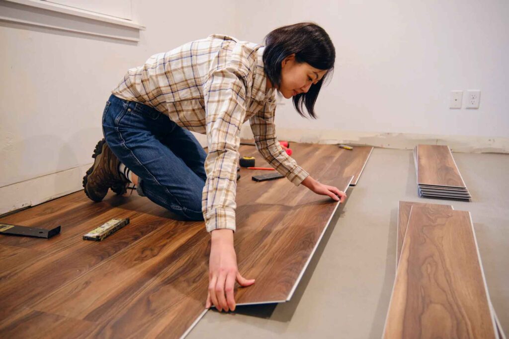 Woman kneeling on the floor installing dark wood laminate flooring planks in a room with tools scattered nearby.