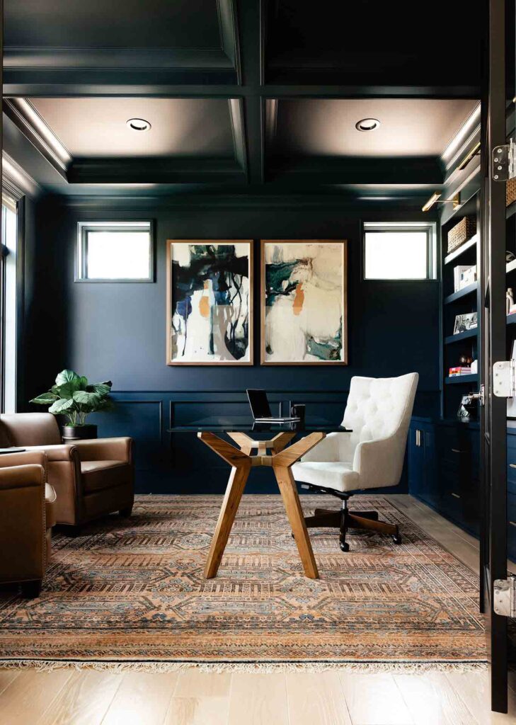 Modern home office with dark navy walls, coffered ceiling, abstract artwork, leather and tufted chairs, wood desk, and patterned area rug on light wood flooring.