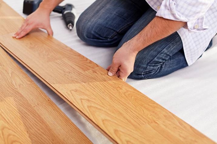 Man kneeling on floor installing wooden laminate planks with a drill tool nearby.