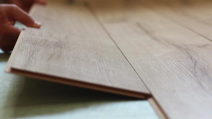 Close-up of hands installing light wood laminate flooring with interlocking planks on a green underlayment.
