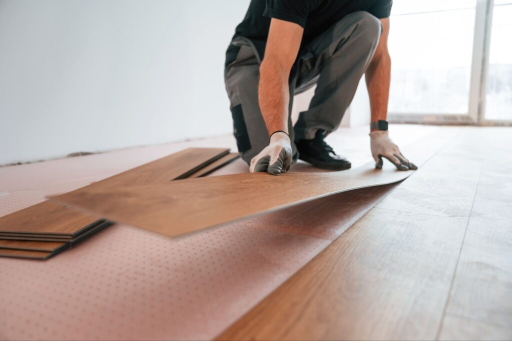 Arbeiter beim Verlegen von SPC-Dielen, Nahaufnahme der Hände beim Verlegen von Vinylfliesen, braune Oberfläche in Holzoptik, rosafarbene Unterlage, Bodenbelag bei der Renovierung eines Hauses, professioneller Verleger kniend, moderne SPC-Dielenverlegung, langlebiges Bodenmaterial, schrittweise Verlegung, lichtdurchfluteter Innenraum.